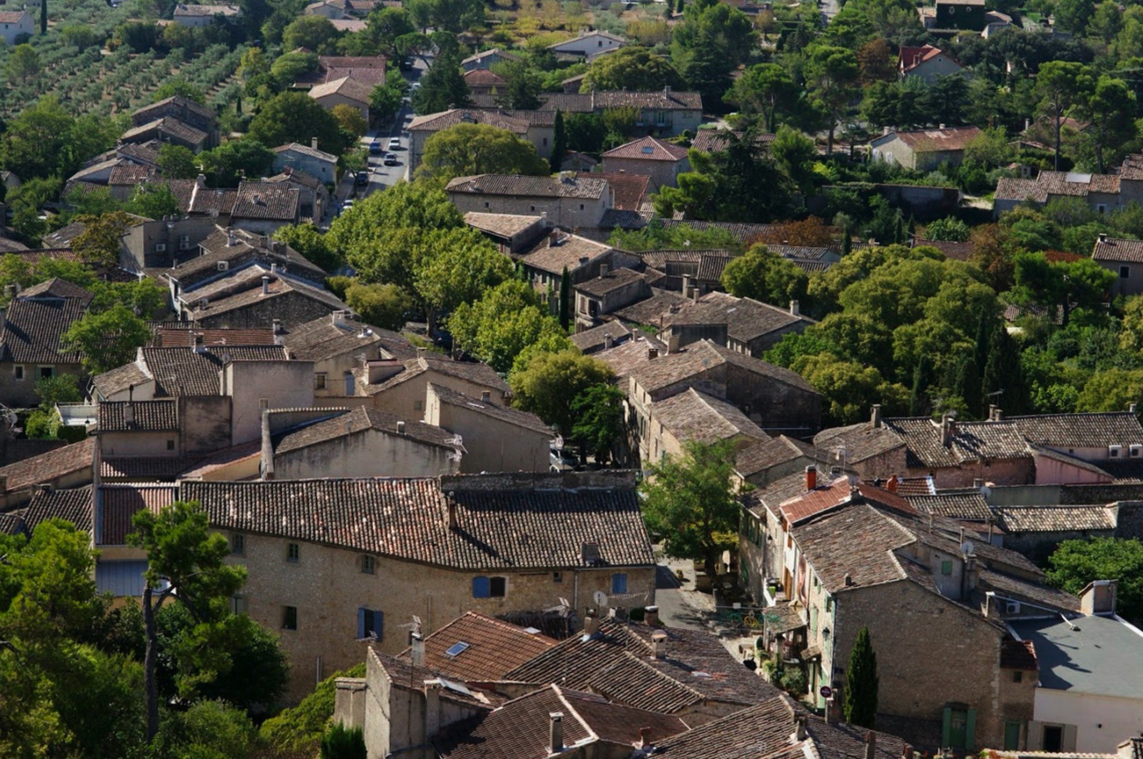 Aerial view of a quaint, old village with tightly packed stone houses and trees scattered throughout, under a clear blue sky.